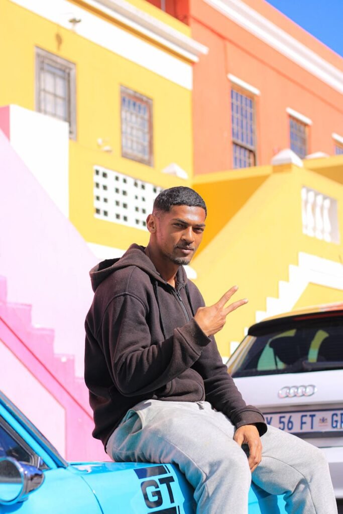 Urban portrait of a man sitting on a car, making a V sign, in front of vibrant buildings.